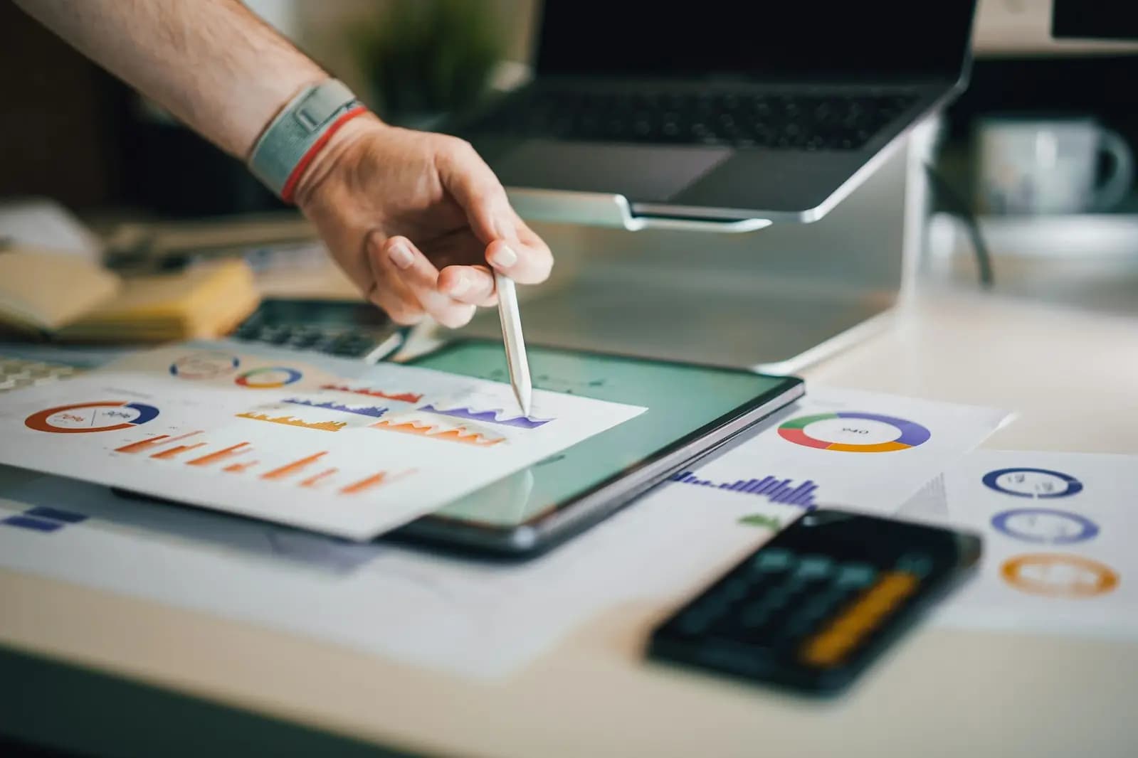 A hand marking up printed charts beside a tablet of reports