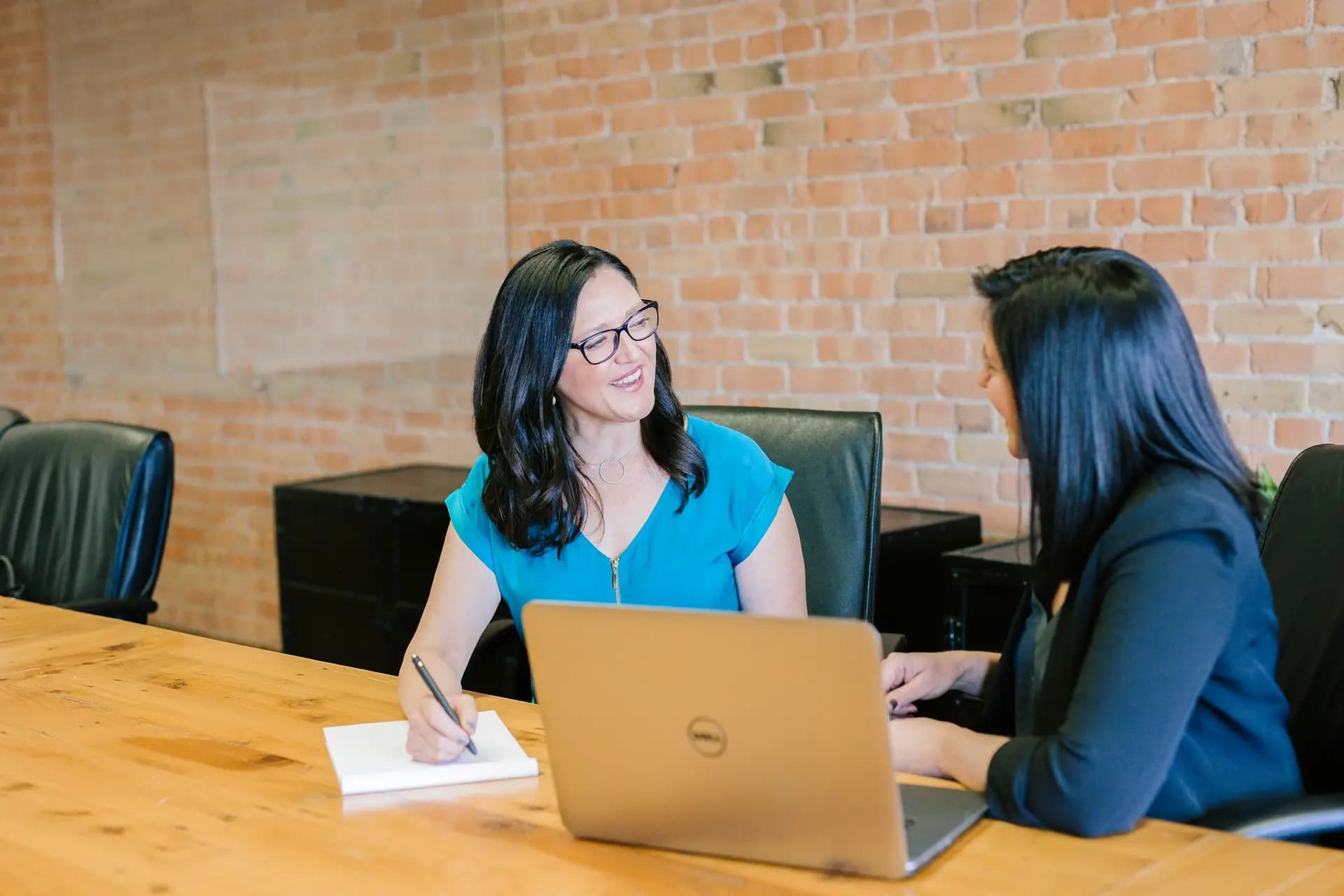 Two colleagues talking across a desk with a laptop in a brick walled office